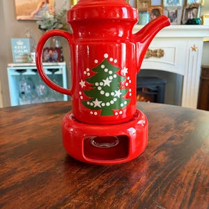 May include: A red ceramic teapot with a matching warmer base. The teapot features a green Christmas tree design with white star and dot accents. The teapot has a handle, a spout, and a lid with a small knob. The warmer base has an opening for a tea light.