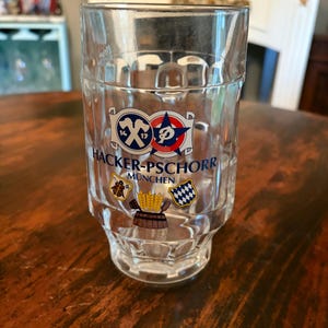 May include: A clear glass beer stein with the words "HACKER-PSCHORR MUNCHEN" printed on the front. The stein has a decorative design with a blue and red logo and other graphics. The stein is sitting on a wooden table.