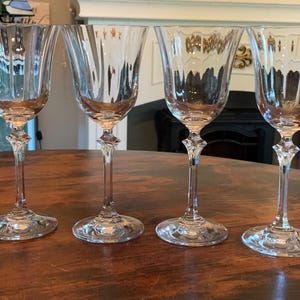 May include: Four clear crystal wine glasses with intricate etched designs. Each glass features a decorative stem and a wide, flared bowl. The glasses are arranged on a dark wooden surface, showcasing their elegant form and clarity.