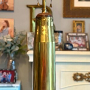 May include: A brass table lamp with a cylindrical base and three light sockets. The lamp sits on a wooden base. The lamp's design is reminiscent of a large bullet casing. The background includes framed pictures.