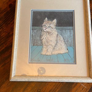 May include: Framed artwork featuring a seated cat illustration. The cat has light brown fur and green eyes, set against a blue and black background. The artwork is in a light-colored wooden frame.