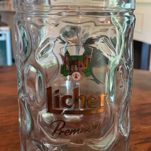 Puede incluir: Jarra de cerveza de vidrio transparente con exterior texturizado. La jarra presenta un logotipo con un diseño de castillo y la palabra "Licher" en letras doradas. También es visible la palabra "Premium". La jarra está sobre una superficie de madera.