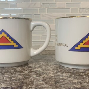 May include: Two white ceramic mugs with gold trim. Each mug features a blue triangle with a yellow and red design. The text "OFFICE OF THE INSPECTOR GENERAL" is printed on the side of each mug.