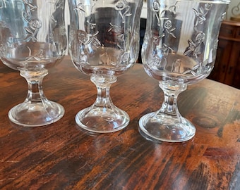 May include: Three clear glass goblets with floral designs. The goblets have a tulip shape with a stem and a round base. The floral pattern is etched into the glass, adding a delicate touch to the glassware.