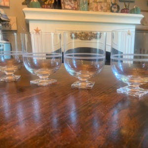 May include: Four clear glass dessert or cocktail glasses with a square base and a wide, rounded bowl. Each glass has a white horizontal band etched around the middle. The glasses are arranged on a dark wooden surface.