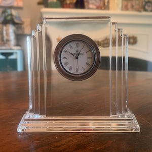 Puede incluir: Un reloj de escritorio transparente de estilo Art Déco con una esfera redonda. La esfera tiene números romanos y la palabra "WATERFORD" impresa. El reloj es de cristal transparente y está sobre una mesa de madera.