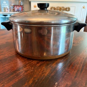 May include: A stainless steel cooking pot with a polished finish, featuring two black plastic side handles and a matching lid with a black knob. The pot has a cylindrical body and a flat base, reflecting light on its metallic surface.
