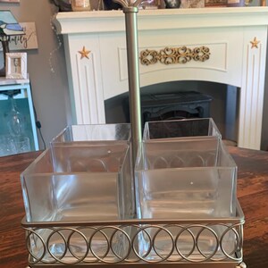 May include: A silver-toned serving caddy with four clear square containers. The caddy has a handle and a decorative base with a repeating circle pattern. The caddy is designed for serving condiments or small items.