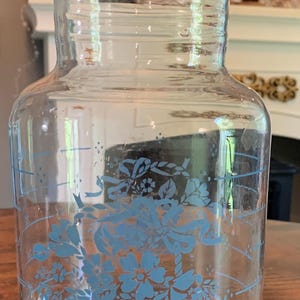 Puede incluir: Jarra de vidrio transparente con boquilla y diseño floral azul. La jarra tiene una base ancha y un cuello estrecho. El diseño floral incluye flores, lazos y hojas. La jarra mide aproximadamente 20 cm de altura.