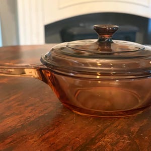 May include: A small, round, amber-colored glass saucepan with a matching lid. The cookware features a single, elongated handle extending from the side and a circular knob on the center of the lid. The glass is translucent and tinted brown.