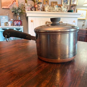 May include: A stainless steel saucepan with a copper bottom and a black handle. The pot has a lid with a black knob. The saucepan is sitting on a dark wooden table. Kitchenware.