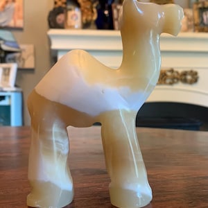 Pode incluir: Estatueta de camelo esculpida em tons de creme e bege. A escultura tem um acabamento liso e polido e está de pé com a cabeça erguida. A forma do camelo é estilizada, com traços arredondados e uma postura graciosa. Um item decorativo.