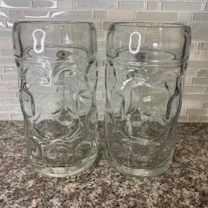 May include: Two clear glass beer steins with a traditional German design. The steins have a rounded bottom and a wide mouth.