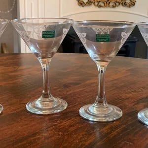 May include: Four clear martini glasses with etched floral designs on the bowl. Each glass has a long stem and a wide, shallow bowl. The glasses are arranged on a dark wooden table. Each glass has a green and white label.