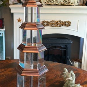 May include: A clear, multi-tiered table lamp with copper-colored accents. The lamp has a pagoda-like design with a silver-colored top and a clear cord. The lamp is on a wooden table.