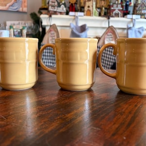 Puede incluir: Tres tazas de cerámica amarilla con un diseño acanalado están dispuestas sobre una mesa de madera oscura. Cada taza tiene un asa curva y una forma cilíndrica. Las tazas están espaciadas uniformemente y parecen nuevas.