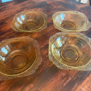 May include: Four amber-colored glass bowls with a decorative floral pattern around the edges. The bowls are oval-shaped with a slightly raised rim and sit on a dark wooden surface. The bowls are likely vintage or antique.