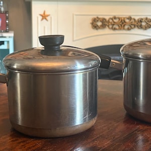 May include: Two stainless steel saucepans with copper-toned bases and black lids. The left saucepan features a long black handle with a metal hanging loop, while the right saucepan has a shorter black handle. Both lids have black knobs.