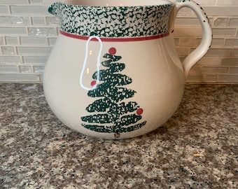 May include: A white ceramic pitcher with a green and red Christmas tree design. The pitcher has a speckled green glaze and a red band around the top.