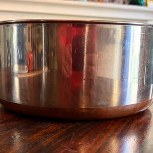 以下が含まれることがあります： 銅色のベースと縁が付いたステンレス製の調理鍋。鍋には2つの黒いハンドルがあり、周囲の環境を反射しています。鍋は木製のテーブルの上に置かれています。