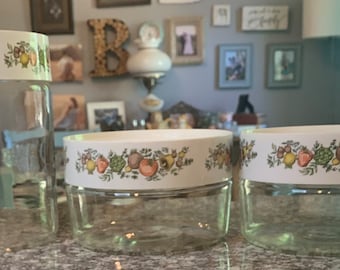 Pode incluir: Três potes de vidro transparente com tampas brancas. As tampas são decoradas com um padrão colorido de frutas e vegetais. Os potes são de tamanhos diferentes, adequados para armazenamento na cozinha.