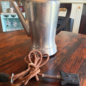 May include: Vintage silver-toned electric coffee pot with a dark brown handle and a clear glass lid. The pot has a long spout and a decorative floral design. A brown, fabric-covered power cord with a black plug is connected to the pot.