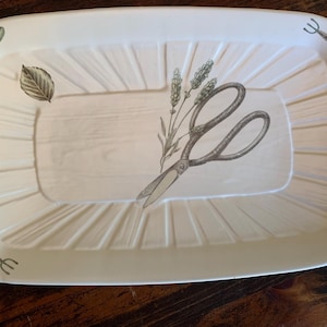 Puede incluir: Bandeja rectangular de color crema con temática de jardín. La bandeja presenta una ilustración central de tijeras y lavanda, con imágenes más pequeñas de herramientas de jardinería, hojas y abejas alrededor del borde. La bandeja tiene un diseño acanalado.