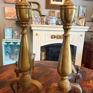 May include: Pair of antique brass and gold-toned andirons. Each andiron has a decorative top, a long, tapered body, and curved feet. The andirons are displayed on a dark wooden table in front of a fireplace.