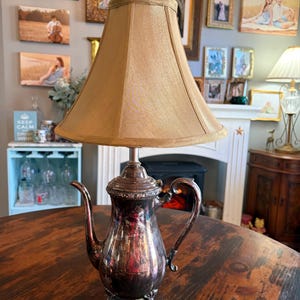 Może przedstawiać: Lampa w stylu vintage z podstawą w kształcie srebrnego czajnika i beżowym abażurem. Podstawa czajnika ma uchwyt i wylewkę, a abażur ma klasyczny stożkowy kształt. Lampa stoi na ciemnym drewnianym stole.