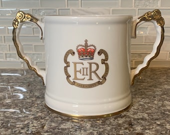 May include: White ceramic mug with gold handles and a gold trim. The mug features a royal crest with a crown, the letters "EIIR", and the text "The Queen's Silver Jubilee 1952-1977".