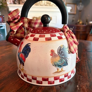 May include: A decorative white and red tea kettle with a black handle and a red lid. The kettle features rooster illustrations and a red and yellow plaid ribbon. The base and top rim have a red and white checkered pattern.