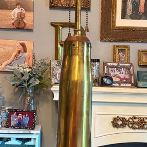 May include: A tall, brass-colored lamp with a cylindrical base and a wooden square base. The lamp has a long, slender neck and a bulb socket at the top. The lamp is on a dark wooden table.