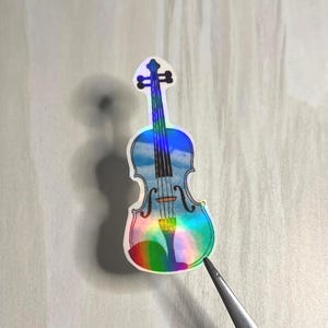May include: A holographic violin-shaped sticker. The body of the violin features a blue sky and cloud design, with rainbow hues around the edges. The strings and details are black.