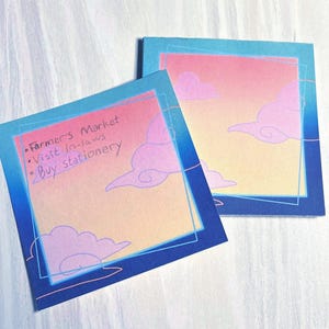 Könnte beinhalten: Zwei quadratische Haftnotizen mit einem Farbverlauf von Blau zu Orange und Pink. Das Design zeigt stilisierte rosa Wolken und einen dünnen blauen Rand. Eine Notiz enthält den handschriftlichen Text: Farmer's Market, Visit in-laws, Buy stationery.