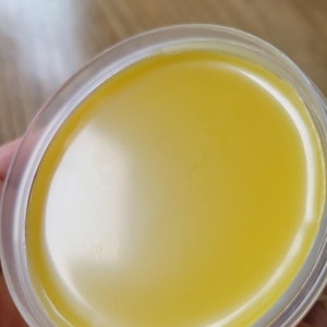 May include: A close-up view of a small, clear plastic container filled with a smooth, bright yellow substance. The surface of the substance is reflective, with a slight sheen. The container is round and appears to be partially filled.