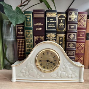 May include: A cream-colored ceramic mantel clock with a gold-tone rim and Roman numeral dial. The clock features embossed fruit motifs, including apples and pears, on the front. The word Lenox is printed in the center of the clock face.