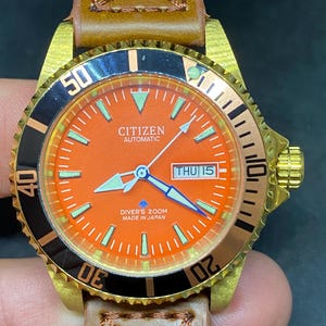 Peut inclure: Une montre de plongée automatique Citizen avec un cadran orange, une lunette noire et dorée, et un bracelet en cuir marron. La montre affiche la date et possède des aiguilles et des index luminescents. Elle est marquée "Diver's 200M Made in Japan".