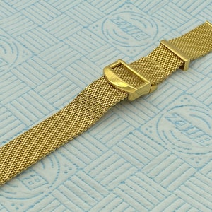 May include: A gold-coloured mesh watch strap with a buckle. The strap is made of a fine, woven metal material. The buckle has the letters "IWC" engraved on it. The strap is on a light blue patterned surface.
