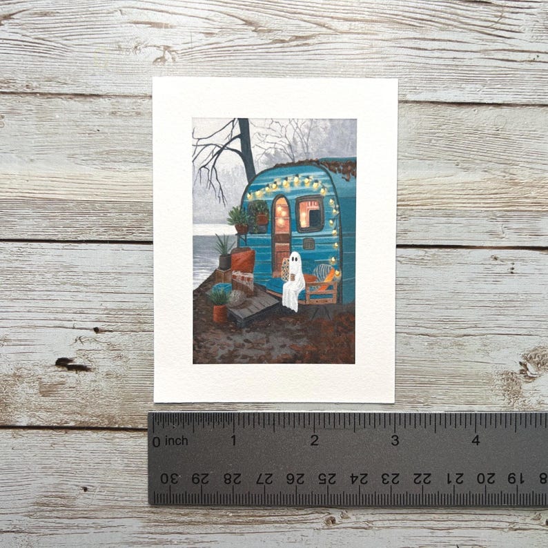 May include: A watercolour painting of a teal camper decorated with fairy lights. A ghost figure sits on a chair, holding a mug. The scene is set near a body of water with trees in the background. The artwork is on white paper, with a ruler showing inches.