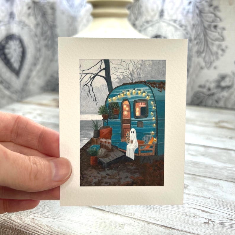May include: A watercolour painting of a teal camper van decorated with fairy lights, a ghost, and autumn leaves. The artwork is on cream paper and held by a hand. The camper door is open, and the interior is illuminated.