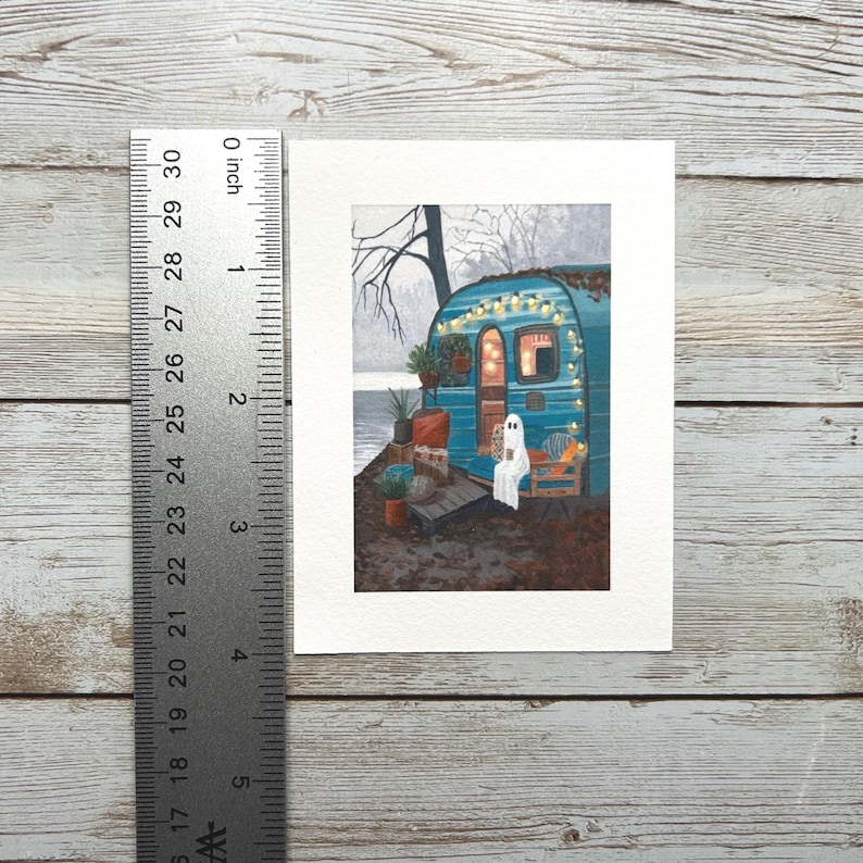 May include: A watercolour painting of a teal camper decorated with fairy lights. A ghost figure sits on a chair on the porch, holding a cup. The artwork is on a white background, with a ruler showing a measurement of approximately 13 cm.