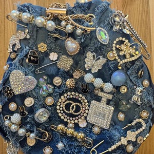 Op de afbeelding: Een denim tas versierd met verschillende versieringen, waaronder strass, parels en metalen bedels. De tas heeft een verweerde look en heeft een verscheidenheid aan vormen en ontwerpen, waaronder harten, vlinders en sterren.