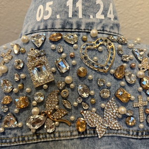 May include: A light blue denim jacket with a white embroidered date "05.11.24" on the collar. The jacket is decorated with rhinestones, pearls, and gold accents, including a heart, a butterfly, and a cross.