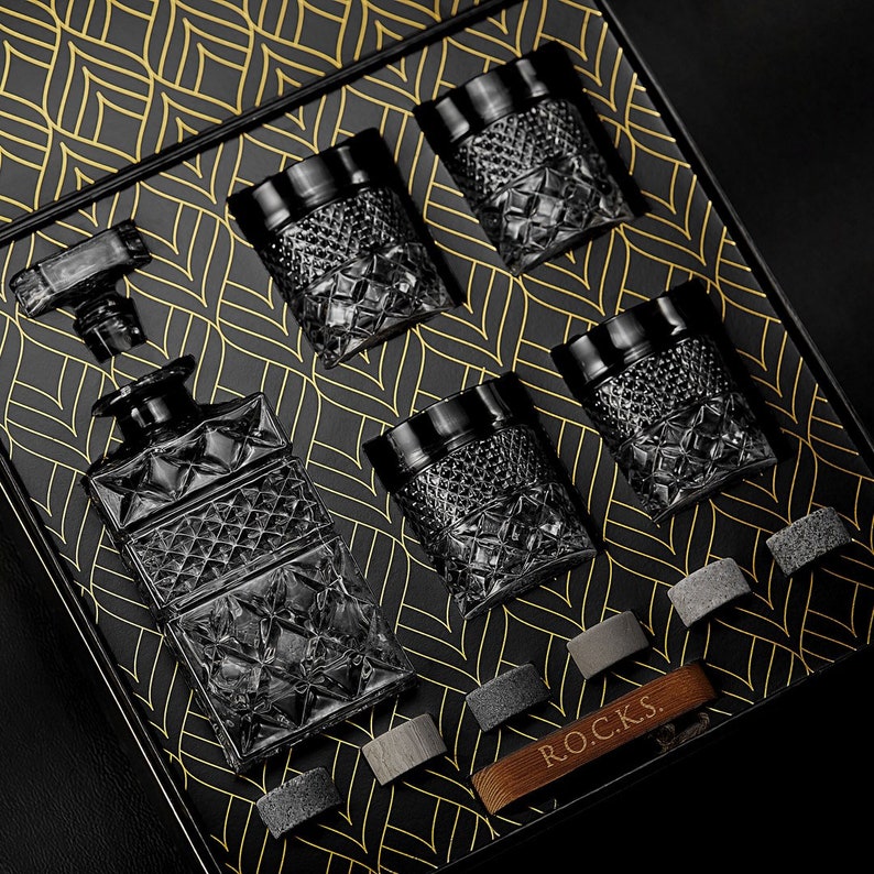May include: A set of six black glass whiskey tumblers with a geometric pattern. The tumblers are arranged in a black box with a gold patterned design. The box also includes a black glass decanter with a geometric pattern. The box has the word "ROCKS" printed on it.