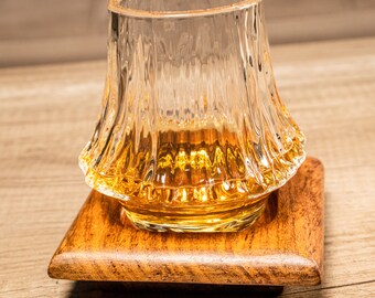 May include: A clear, textured glass with a golden liquid inside, resting on a small, square wooden coaster. The glass has a unique, ridged design, and the coaster has a warm, brown finish. The drinkware is set against a light wood grain background.