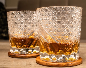 May include: Two clear glass tumblers with a diamond pattern, filled with amber liquid, sitting on wooden coasters.
