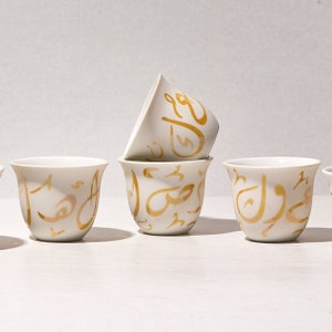 May include: Set of four white ceramic coffee cups with gold Arabic calligraphy designs. The cups are arranged in a row on a white surface.