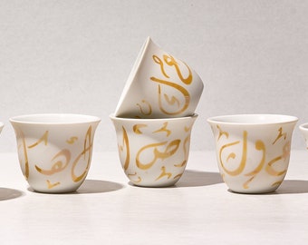 May include: Set of four white ceramic coffee cups with gold Arabic calligraphy designs. The cups are arranged in a row on a white surface.