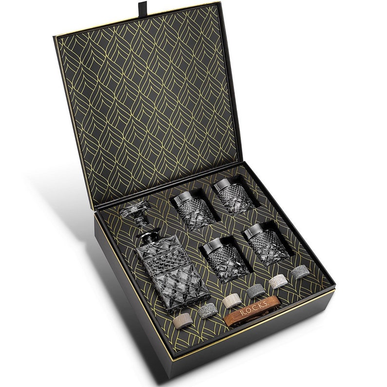 May include: A black gift box with a gold patterned lid is open to reveal a crystal decanter and six crystal glasses. The box also includes a set of whiskey stones and a leather tag that says "Rocks".
