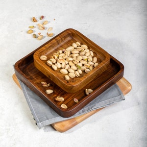 May include: Two wooden square serving trays, one nested inside the other, with a pile of pistachios in the smaller tray. The trays are made of dark wood with a natural grain pattern.
